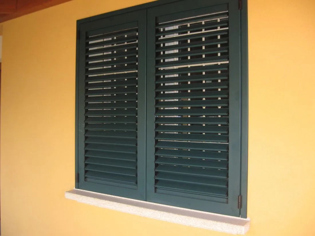 Persiana verde a due ante con lamelle orientabili aperte, su una facciata gialla.