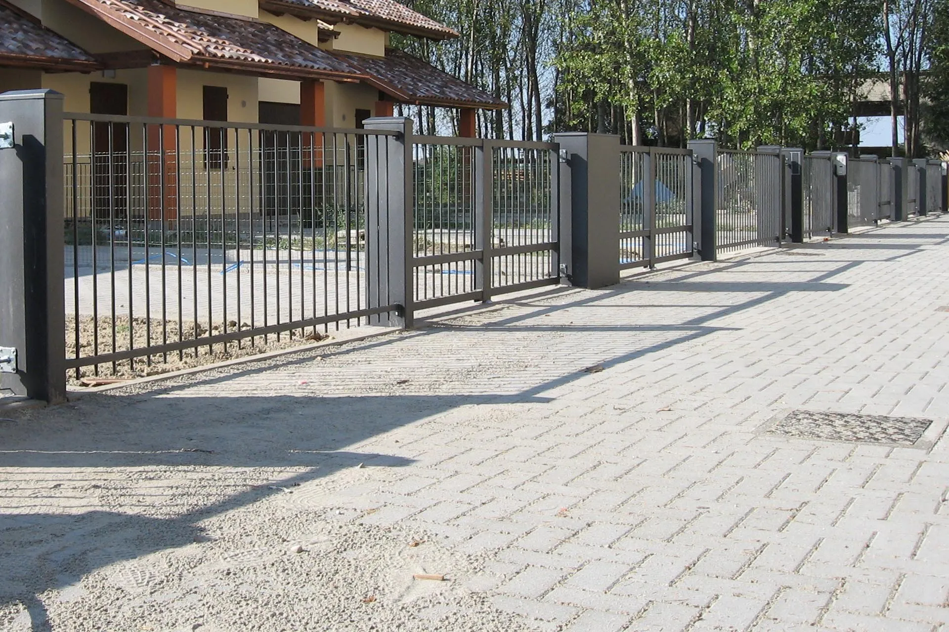 Recinzione in ferro grigio con pilastri e cancelli lungo un viale, davanti a un complesso di villette.