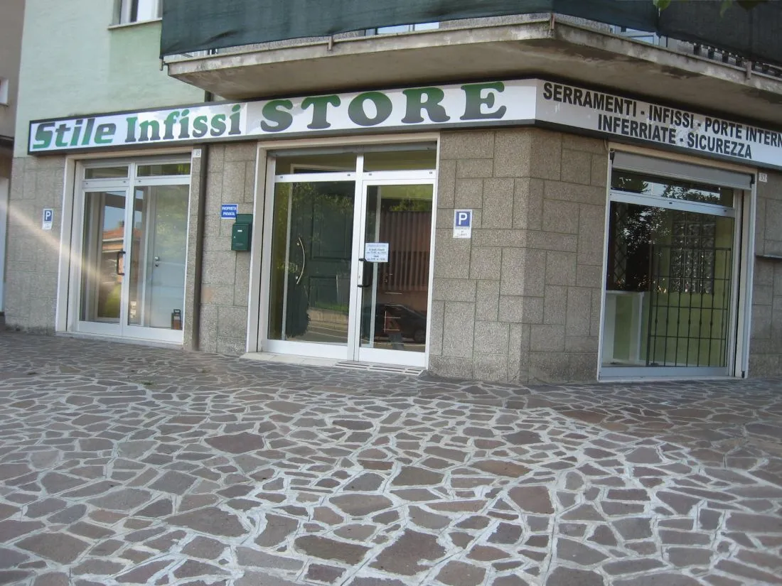 Negozio d'angolo con l'insegna Stile Infissi Store e tre vetrine, visto dalla piazza lastricata.
